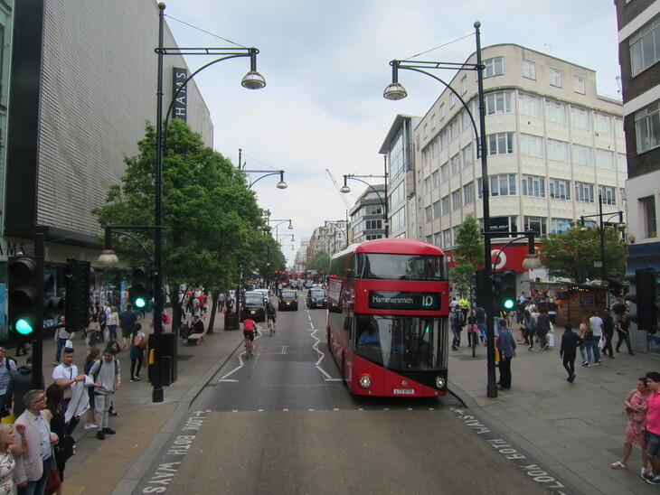 A red double-decker bus travels down a busy London street lined with pedestrians, shops, and tall buildings under a cloudy sky.