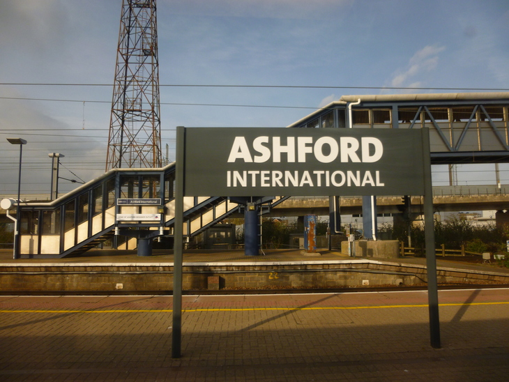 A large sign reading "ASHFORD INTERNATIONAL" stands on a train platform, with a pedestrian bridge and a tall metal lattice tower in the background.