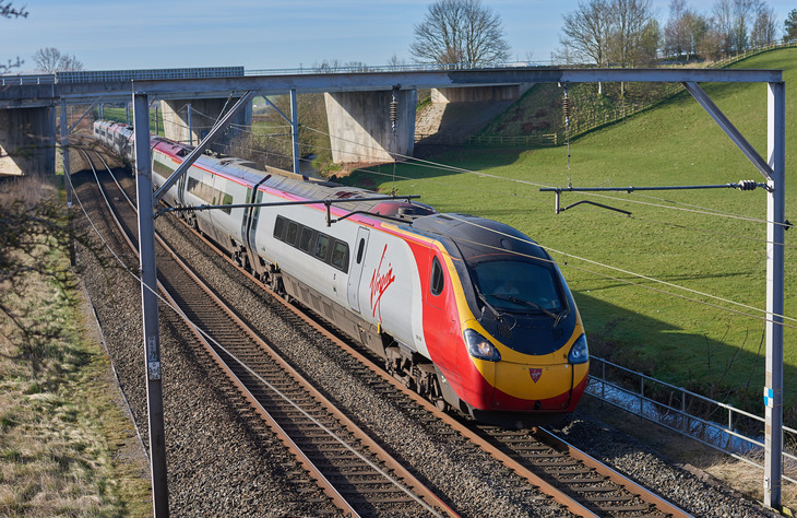 A high-speed Virgin Trains Pendolino travels along a curved track under a concrete bridge and overhead power lines, set against a backdrop of green hills.