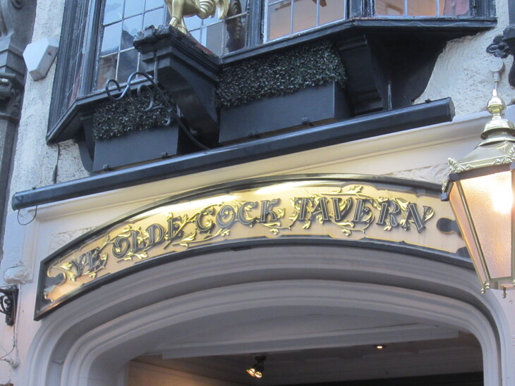 The ornate, gold-lettered sign for Ye Olde Cock Tavern above an arched stone entrance, with a traditional lantern to the right and a bay window above.