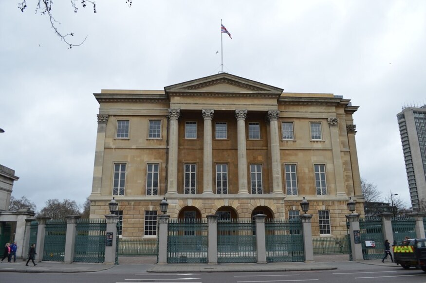 A large, three-story neoclassical stone building with a central portico featuring four Corinthian columns and a triangular pediment, flying a Union Jack flag from the roof. The building is situated behind a green iron fence with stone pillars on a city street.