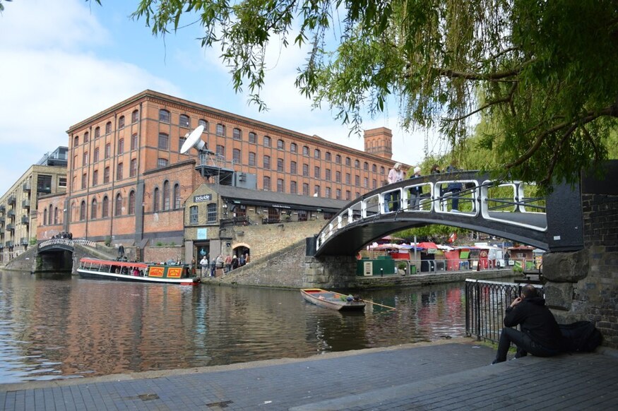 A scenic view of Camden Lock in London, featuring a brick industrial building, a black arched pedestrian bridge over the canal, and a tour boat on the water. A person sits on the paved bank in the foreground, looking out at the canal.