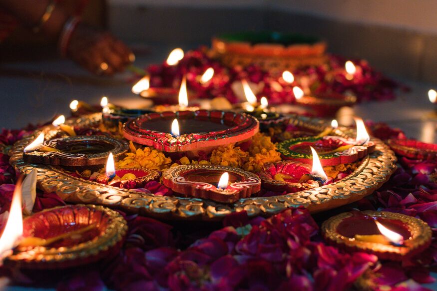 A close-up shot of several lit clay oil lamps, or diyas, arranged on a decorative golden platter and surrounded by red rose petals for a festive celebration.