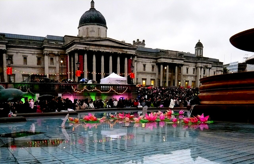 A low-angle view of the National Gallery in London's Trafalgar Square at dusk, with pink lotus flower lanterns floating in a large fountain in the foreground. A crowd of people is gathered behind the fountain, and the building's reflection is visible on the water's surface.