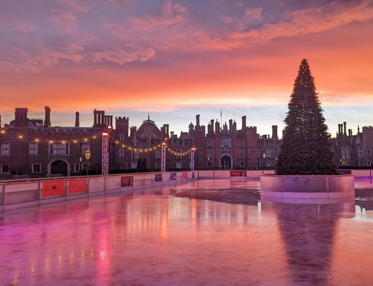 Where to go ice skating in London Christmas 2026: An empty ice rink with a Christmas tree at the centre, in front of Hampton Court Palace beneath an orange and pink sunrise