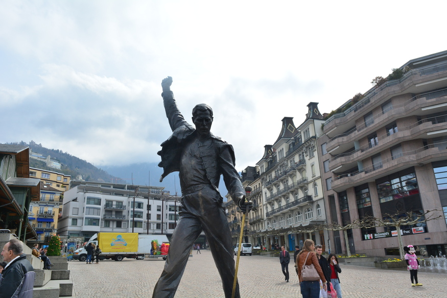 A bronze statue of Freddie Mercury stands in a town square in Montreux, Switzerland, with his right arm raised in a fist and his left hand holding a microphone stand. Buildings and a cloudy sky are in the background.