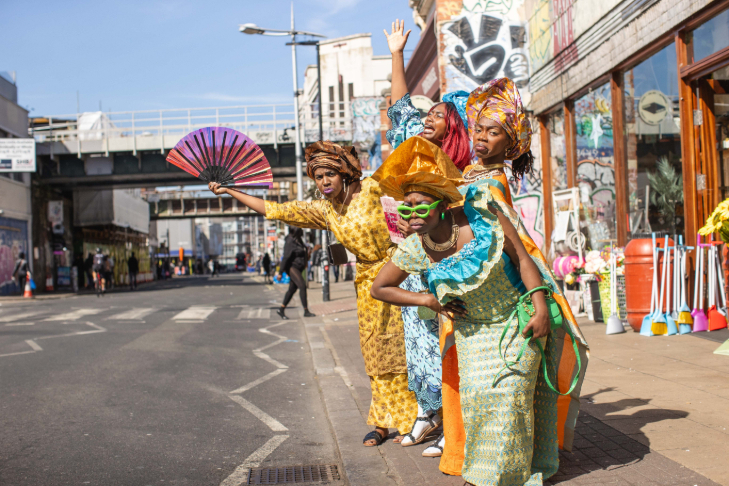 A group of women standing on the edge of the pavement in Peckham in colourful costumes, with one holding a fan out into the road as if hailing a bus