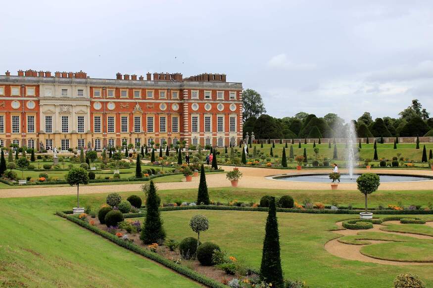 A wide view of the ornate red-brick facade of Hampton Court Palace overlooking formal gardens with manicured lawns, topiary trees, and a central fountain.