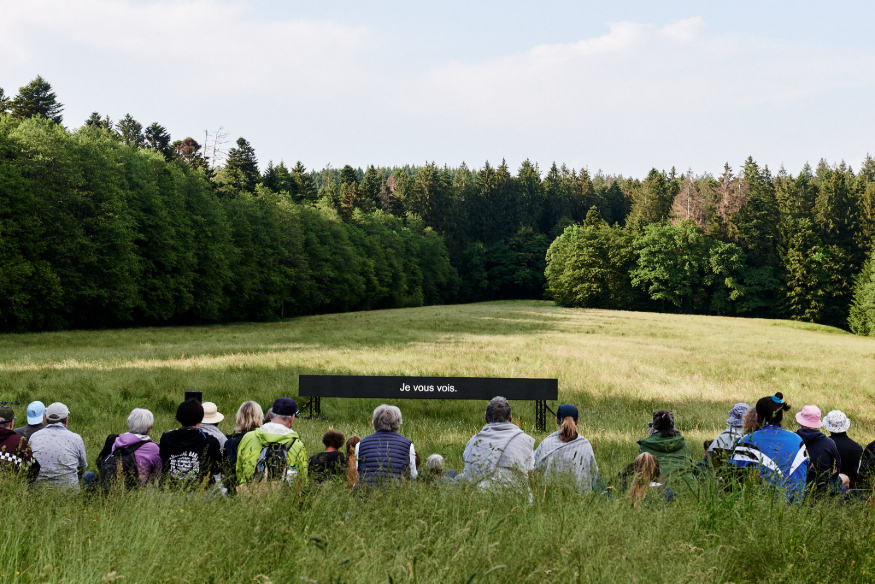 A group of people sitting in grass on a hilly field, looking towards a sign.