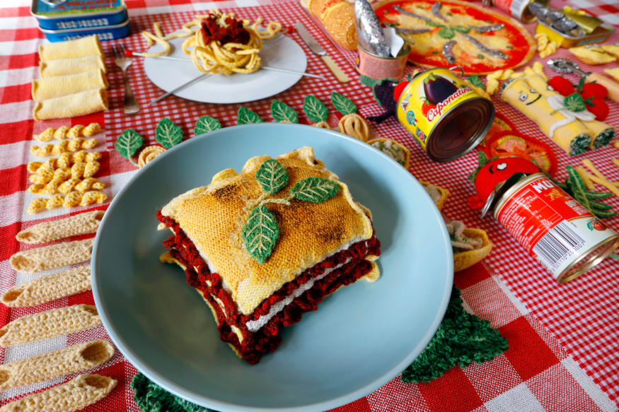 A lasagne made entirely of wool plated up on a table, surrounded by knitted food items including penne pasta and sliced tomatoes