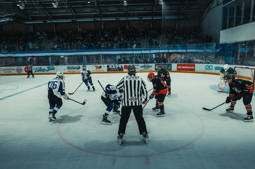 A wide shot of an indoor ice hockey game during a face-off, with a referee in a striped jersey standing between two opposing players in blue and black uniforms while spectators watch from the stands.