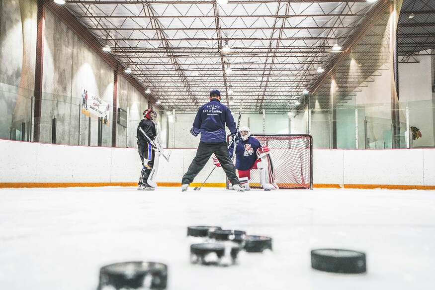A low-angle shot of a hockey goalie training session on an indoor ice rink, with several pucks in the foreground and a coach instructing two goalies in front of a net.