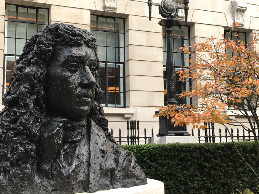 A large, dark bronze bust of a man with long, curly hair stands in the foreground, set against a light-colored stone building with large windows, a black iron fence, and a tree with orange autumn leaves.