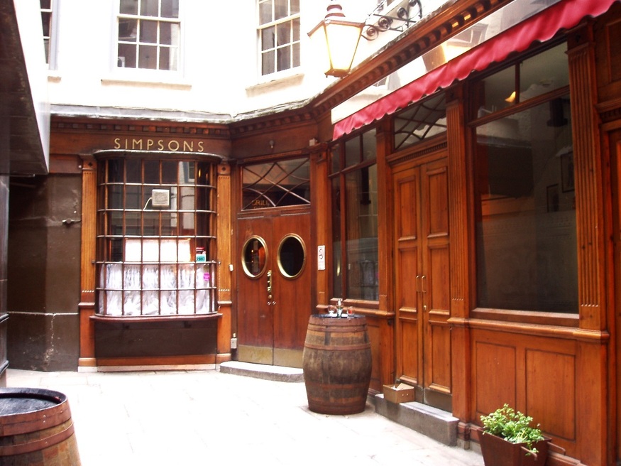 The wooden exterior of Simpsons Tavern in London, featuring a curved bay window, double doors with porthole windows, and a large wooden barrel in a narrow alleyway.