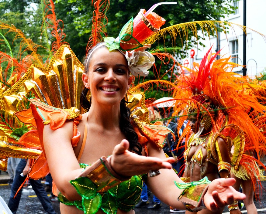 Things to do in London August bank holiday weekend 2026:  A smiling woman in a vibrant carnival costume with gold, orange, and green accents, featuring a large feathered headpiece and a decorative cup on her head. She extends her hands toward the camera, with another performer in a similar orange feathered costume visible in the background.
