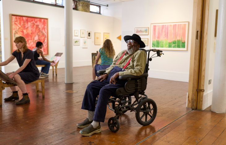 What's on in London today: artist Frank Bowling sitting in a wheelchair in the centre of an art gallery, with other artists working in the background