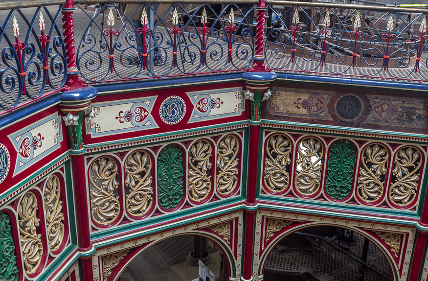 Intricate Victorian cast-iron metalwork featuring ornate red, green, and gold scrollwork panels and a decorative blue railing with gold-tipped finials.