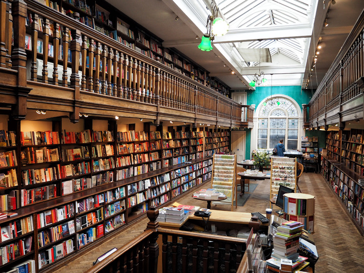 Interior of a multi-level bookstore featuring floor-to-ceiling wooden bookshelves, a mezzanine balcony with a carved railing, and a large skylight illuminating the center aisle.