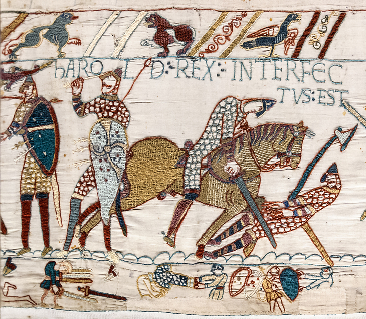 A section of the Bayeux Tapestry depicting the death of King Harold at the Battle of Hastings. Embroidered figures show a soldier being struck in the eye with an arrow and another being cut down by a Norman knight on horseback. Latin text above reads "HAROLD REX INTERFECTUS EST" (King Harold is killed). The scene includes a decorative border with animals and fallen soldiers.