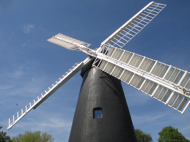 A low-angle shot of a black brick windmill with white sails against a clear blue sky.