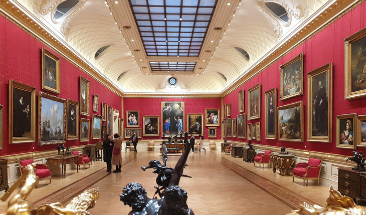 Things to do in London today:  A long, grand art gallery with deep red damask walls densely hung with ornate gold-framed paintings. The room features a vaulted white ceiling with a central skylight, polished wood floors, and red velvet chairs lining the sides. Several visitors walk through the space, and dark bronze sculptures are positioned in the foreground.