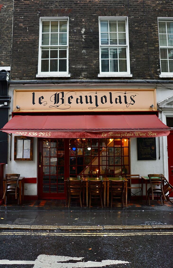 The storefront of Le Beaujolais Wine Bar, a French restaurant with a red awning and outdoor seating on a wet city street.