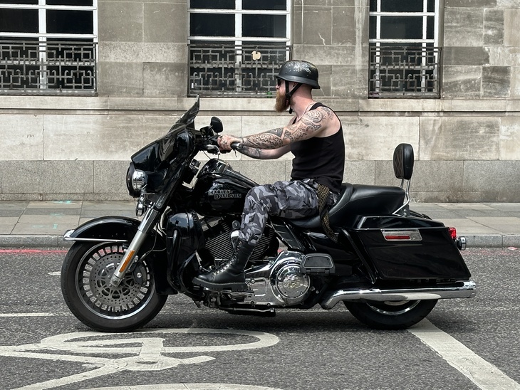 A man with tattoos and a beard rides a black Harley-Davidson motorcycle past a stone building.