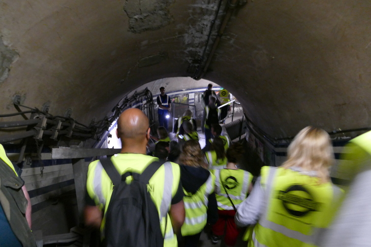 A group of people wearing high-visibility yellow vests walk through a narrow, arched underground tunnel with exposed wiring and pipes along the walls.