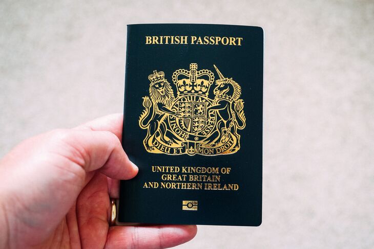 A hand holding a dark blue British passport with gold lettering and the royal coat of arms.