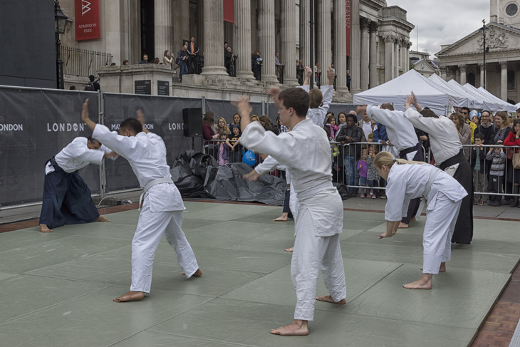 A group of people in white martial arts uniforms perform a demonstration on green mats in a public square, with a crowd and large stone building in the background.