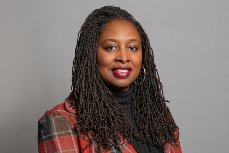 A headshot of Dawn Butler, a Black woman with long dark locs, wearing a red and brown plaid blazer over a black turtleneck, smiling against a plain grey background.