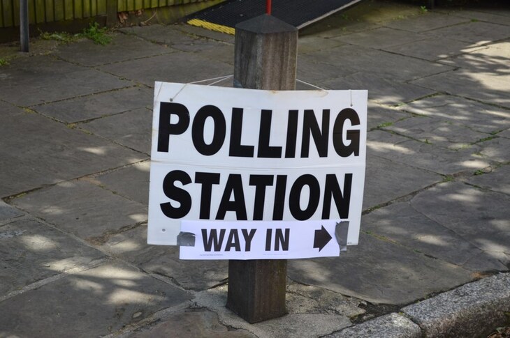 A white sign with black text reading "POLLING STATION" and a smaller taped-on sign below saying "WAY IN" with an arrow pointing right, attached to a wooden post on a stone-paved sidewalk.