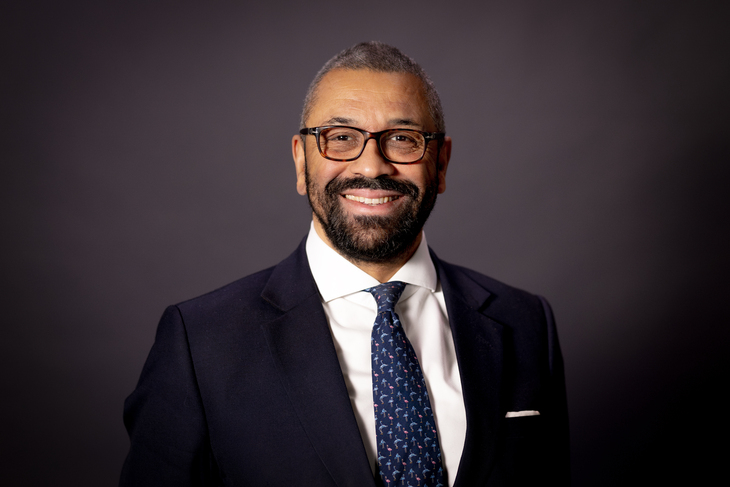 A head-and-shoulders portrait of James Cleverly, a man with short graying hair, glasses, and a beard, smiling in a dark suit, white shirt, and patterned blue tie against a dark gray background.