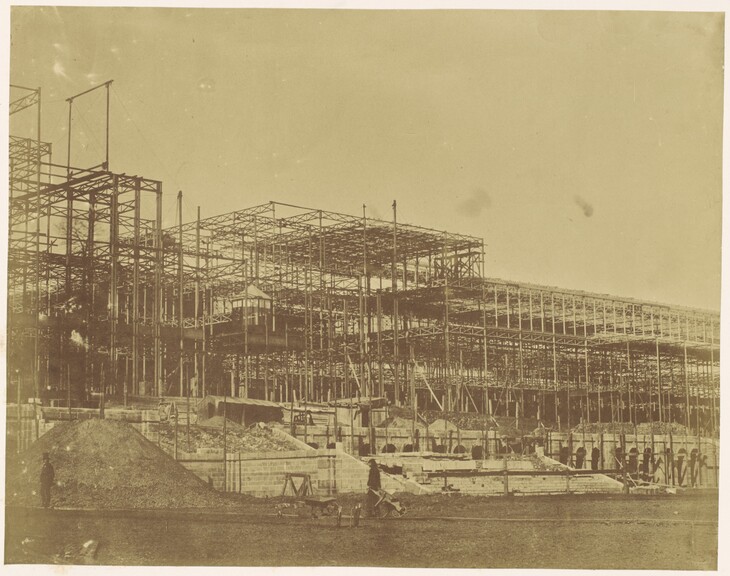 A sepia-toned 19th-century photograph showing the iron and glass framework of the Crystal Palace under construction in Sydenham, with laborers and piles of dirt in the foreground.