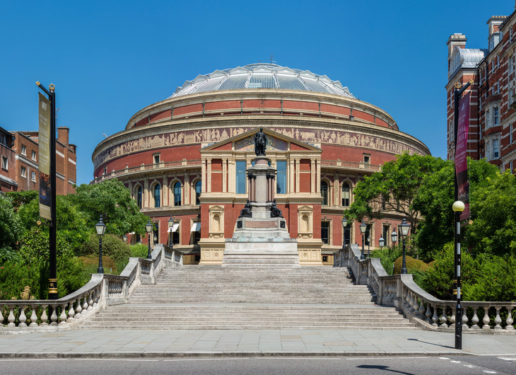 A wide-angle, eye-level shot of the Royal Albert Hall in London, England, featuring its iconic red brick and terracotta circular exterior, glass dome, and the Albert Memorial statue at the top of a grand stone staircase.
