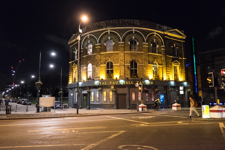 The Royal Vauxhall Tavern, a historic curved brick building, illuminated at night on a London street corner with a cyclist and pedestrian nearby.