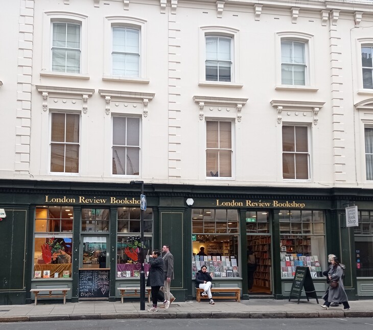 A street-level view of the London Review Bookshop, featuring a dark green storefront with large windows and gold lettering. People are walking by or sitting on benches outside the shop, which is located on the ground floor of a white, multi-story building with classic sash windows.