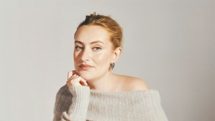 Things to do in London today: A lady in a beige jumper with her hair up resting her head on one hand.