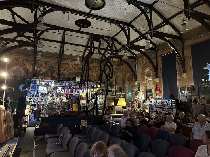 An audience sits in rows of chairs inside a large hall with a high, vaulted wooden ceiling. In the background, a bar area features a neon sign that reads "PALLADIUM