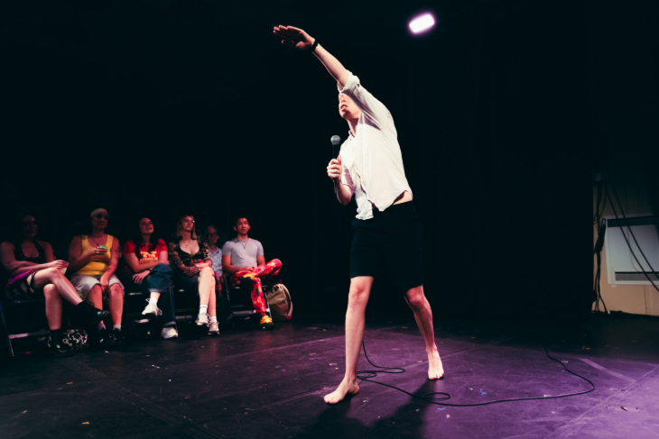 A man in white shirt and black shorts, reaching one arm into the air as he performs on stage in front of an audience