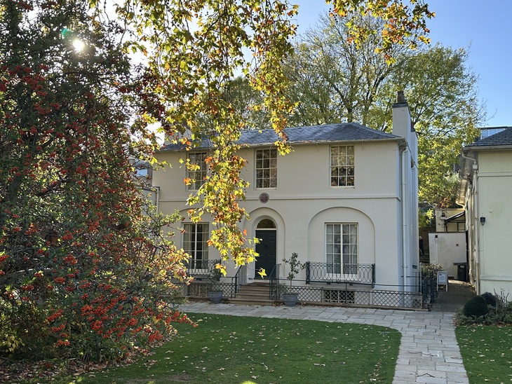 A two-story, cream-colored Regency house with a dark green door and arched windows, partially framed by a tree with red berries and autumn leaves. A stone path leads across a green lawn toward the house under a clear blue sky.