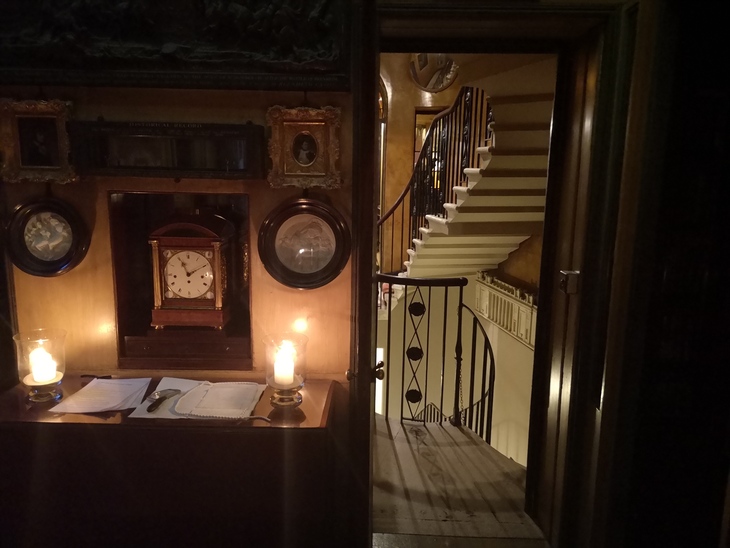 Things to do in London today: A dimly lit, historic interior featuring a wooden sideboard with two lit candles, a mantel clock, and framed portraits. An open doorway reveals a brightly lit, elegant spiral staircase in the background.