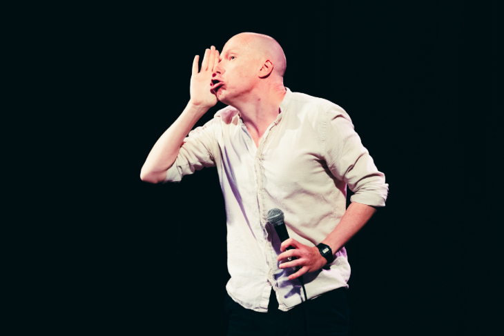 Things to do in London today: Jordan Brookes on stage in a white shirt, holding one hand to his face as if shouting