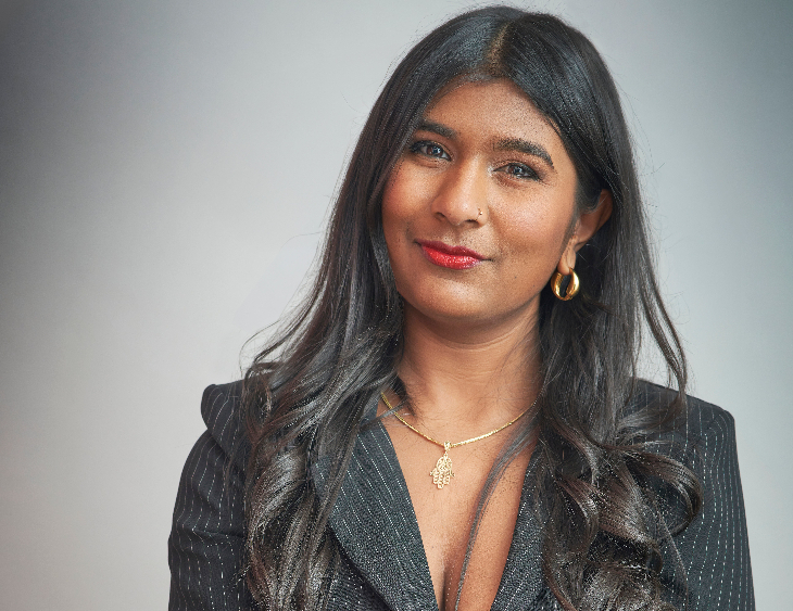 Things to do in London this weekend: Ash Sarkar poses in a pinstripe blazer and gold hamsa necklace against a gray wall.