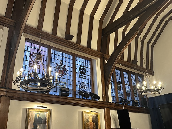 Interior of a historic hall featuring dark timber ceiling beams, stained glass windows with heraldic crests, and two ornate wrought-iron chandeliers. Portraits in gold frames hang on the white walls below the windows.