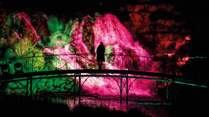 Winter light trails festivals near London Christmas 2026: a person on a footbridge over a lake silhouetted against a waterfall which is illuminated in pink at night