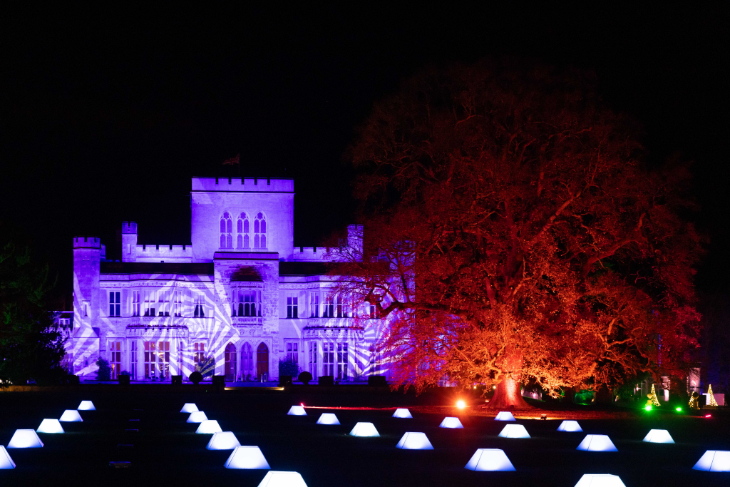 Winter light trails festivals near London Christmas 2026: rows of white lights in front of a manor house at night. A tree alongside the house is lit up in red