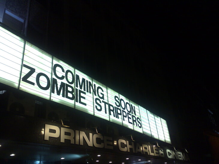A low-angle night shot of the Prince Charles Cinema marquee, which reads "COMING SOON ZOMBIE STRIPPERS" in black block letters against a glowing white background.