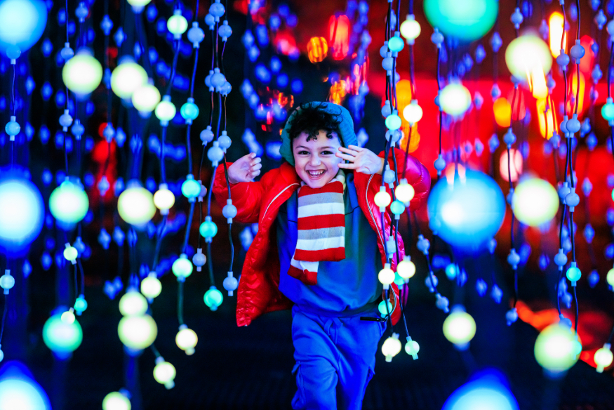 Christmas 2026 best light trails London: a young child running through dangling strings of lights with a smile on their face