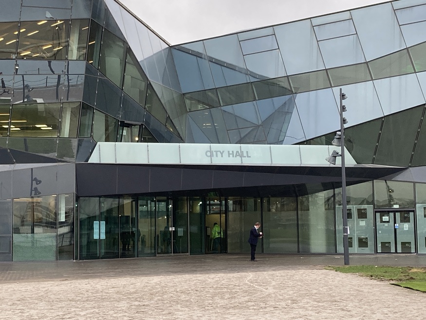 A modern glass building with a sign above the entrance that reads City Hall. A man in a suit stands in front of the glass doors.
